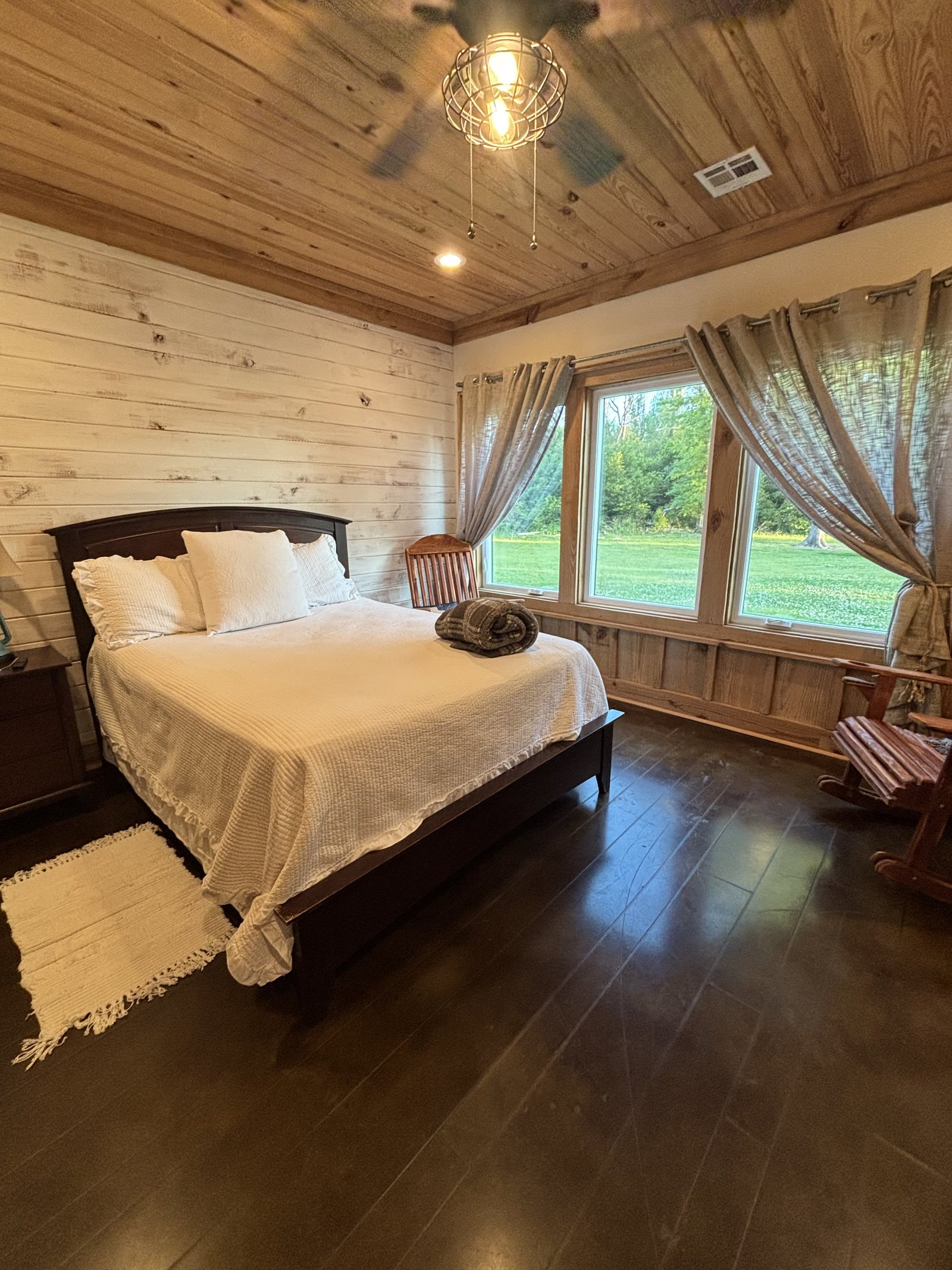 A bedroom in the country house — white linens, shiplap walls, cedar ceiling and a window onto the field