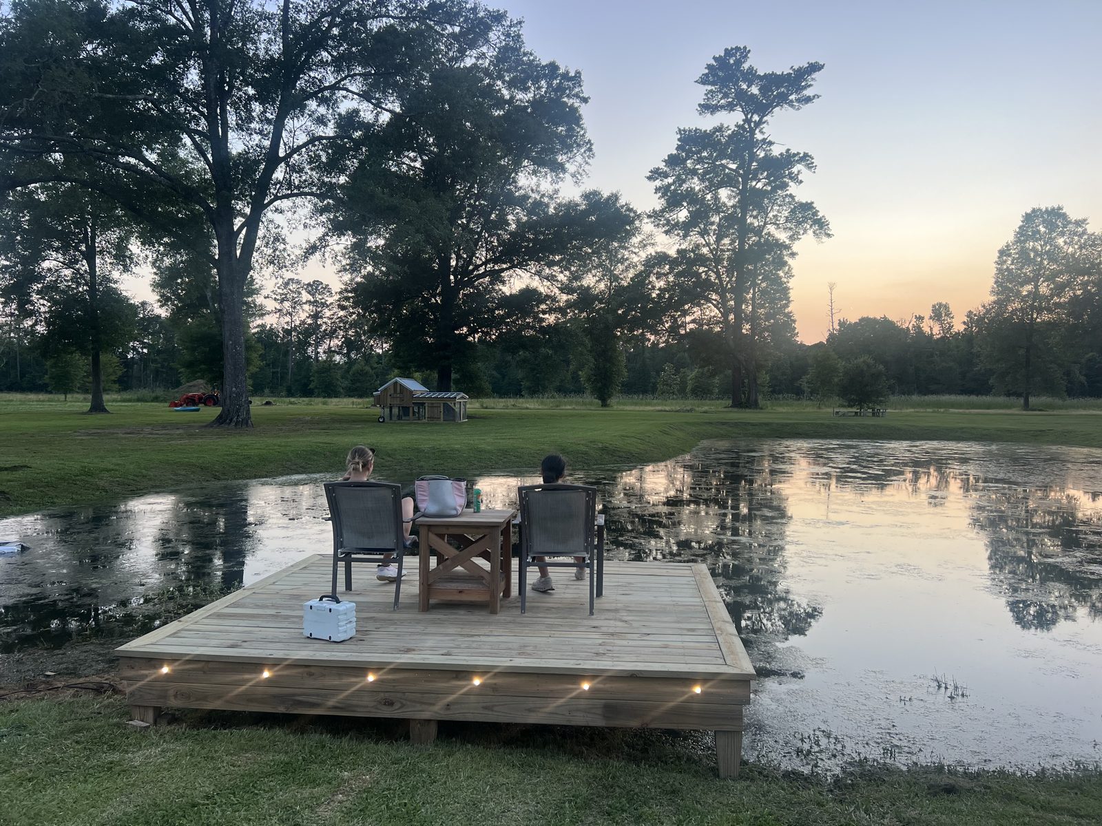 Guests sitting on the lit pond deck watching the sun set behind the tree line